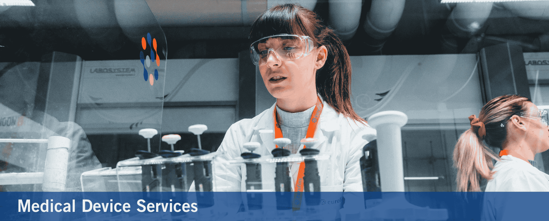 Two female scientists in a laboratory wearing safety goggles and lab coats, working with pipettes and medical devices. The image is related to medical device services, emphasizing precision and expertise in a clinical or research setting
