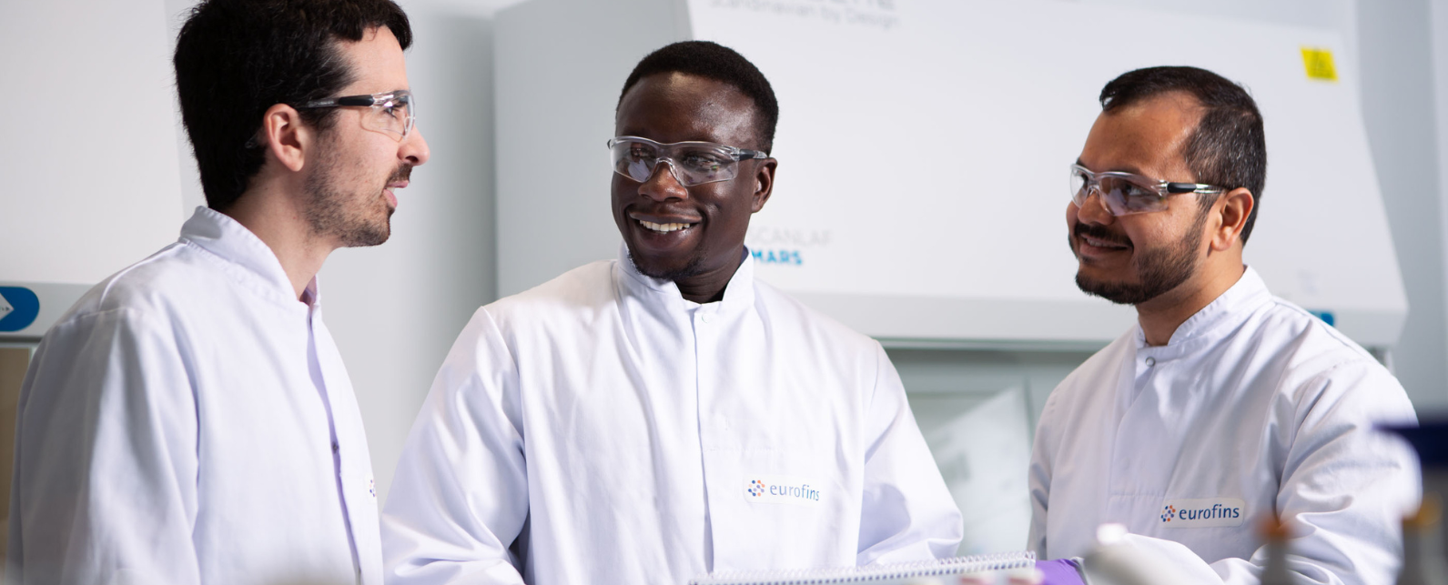 Three consultants in white lab coats and safety glasses talking 