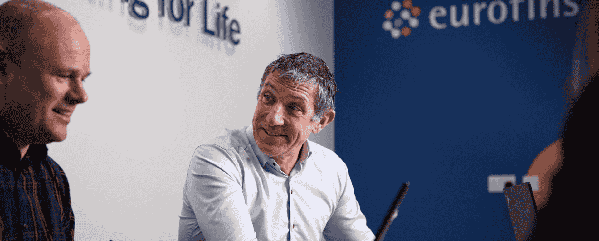 Two consultants engaged in a conversation in a modern office setting, with the Eurofins logo visible in the background. The image captures a collaborative business environment focused on medical devices