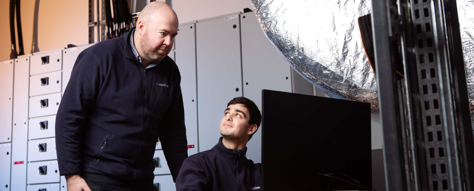 a guy in front of a computer wearing a dark jacket. Behind him there is another person wathing the screen of the computer.