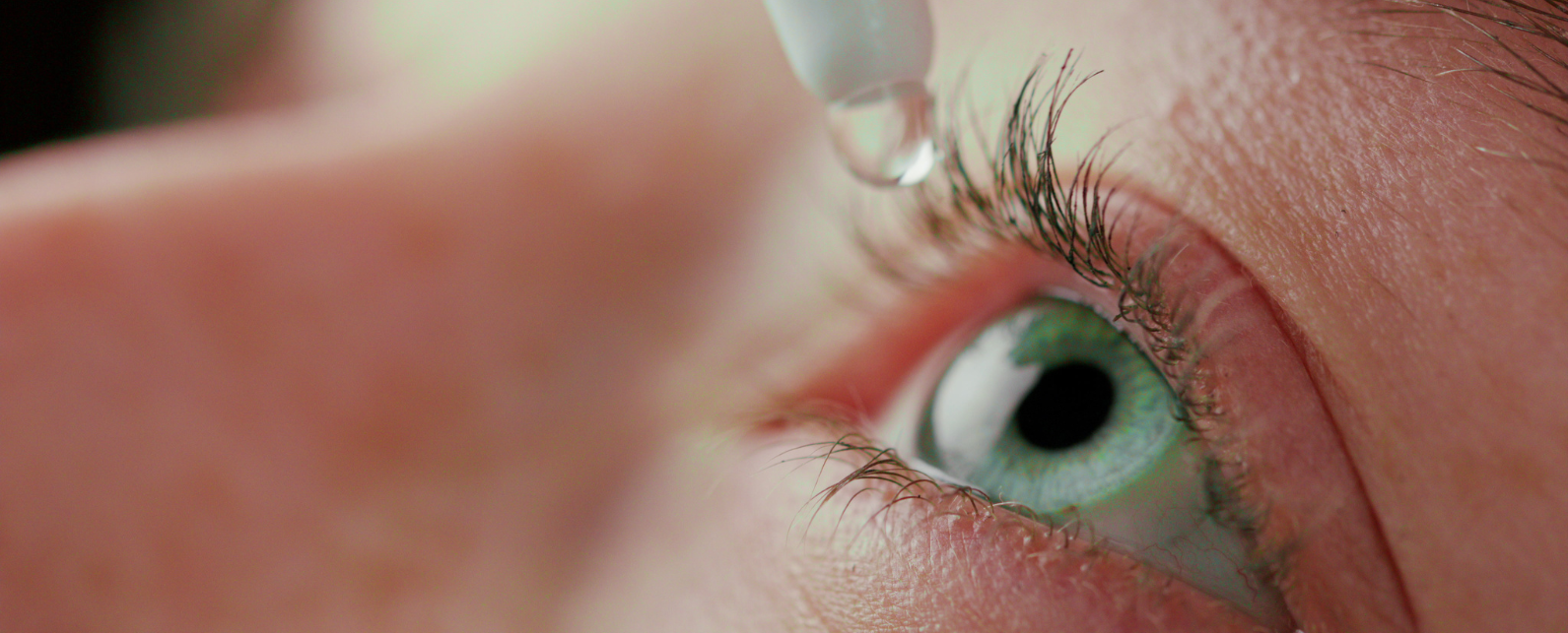 A tip of a white eye drop bottle with an eye drop above a agreen eye