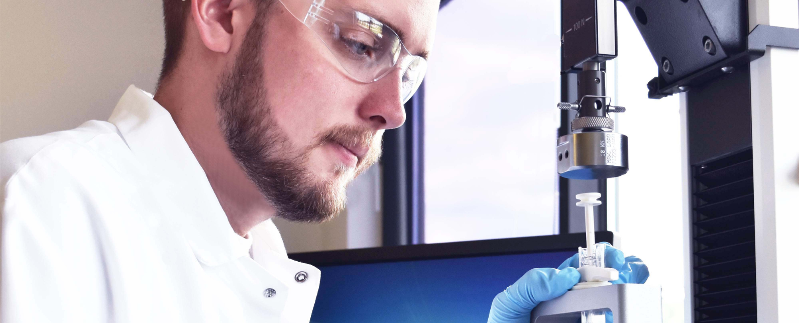 A man in a lab coat with lab glasses uses an Instron machine to test a syringe