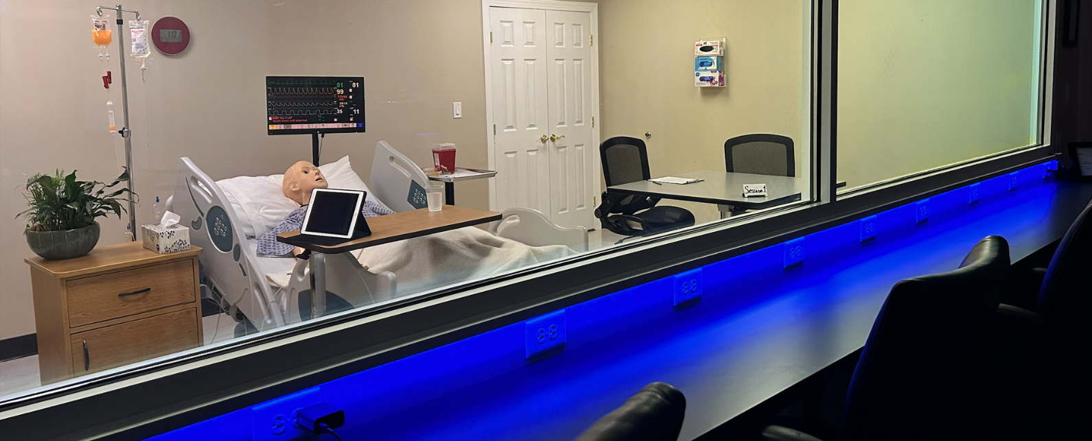 Chairs next to a window to view a lab simulation of a mannequin in a hospital bed with a night stand and IV pole
