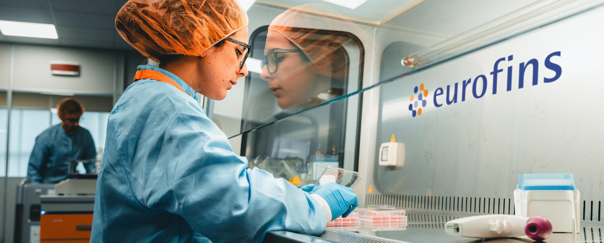 woman in a laboratory wearing a lab coat, hair cover, and gloves conducting cytotoxicity testing