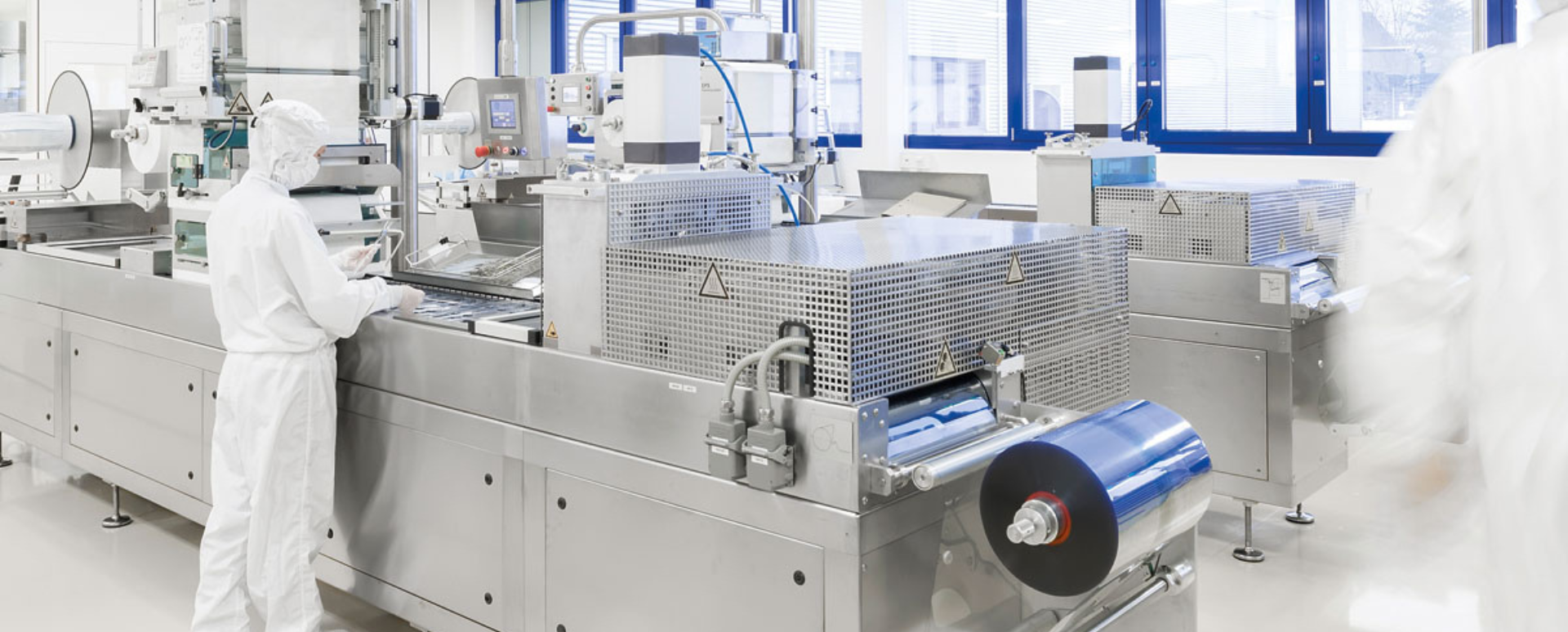 Zeigt einen Produktionsmitarbeiter in einem Reinraum-Overall, der neben einer Blisterproduktionsmaschine steht. 